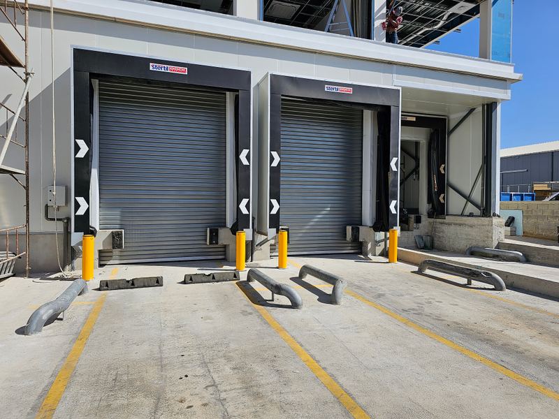 Two Stertil Dock Products loading dock bays with closed roller shutter doors, yellow bollards, and wheel guides.