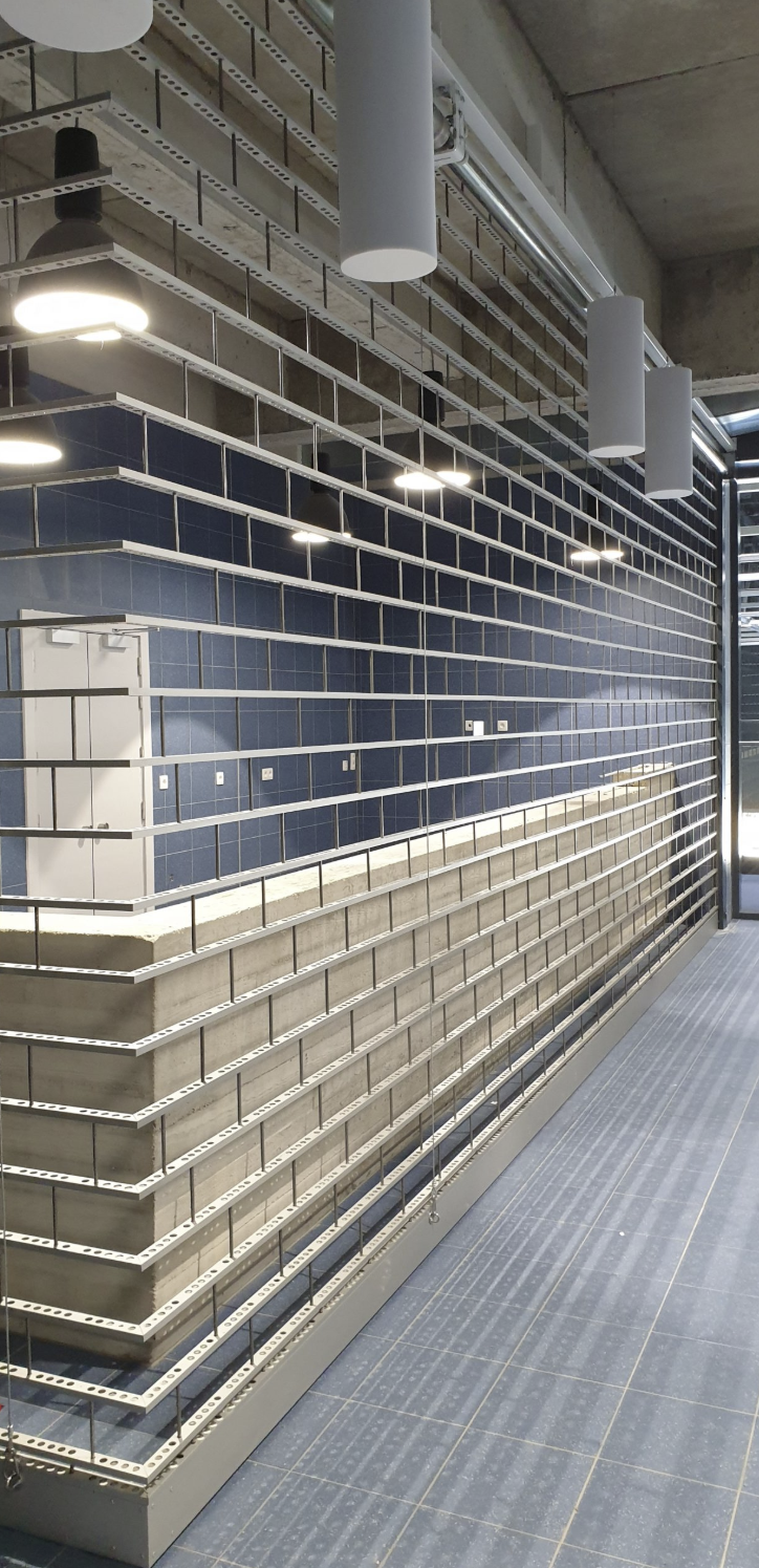 Industrial shelving system with perforated metal channels and concrete base, blue tiled wall, and ceiling lights.
